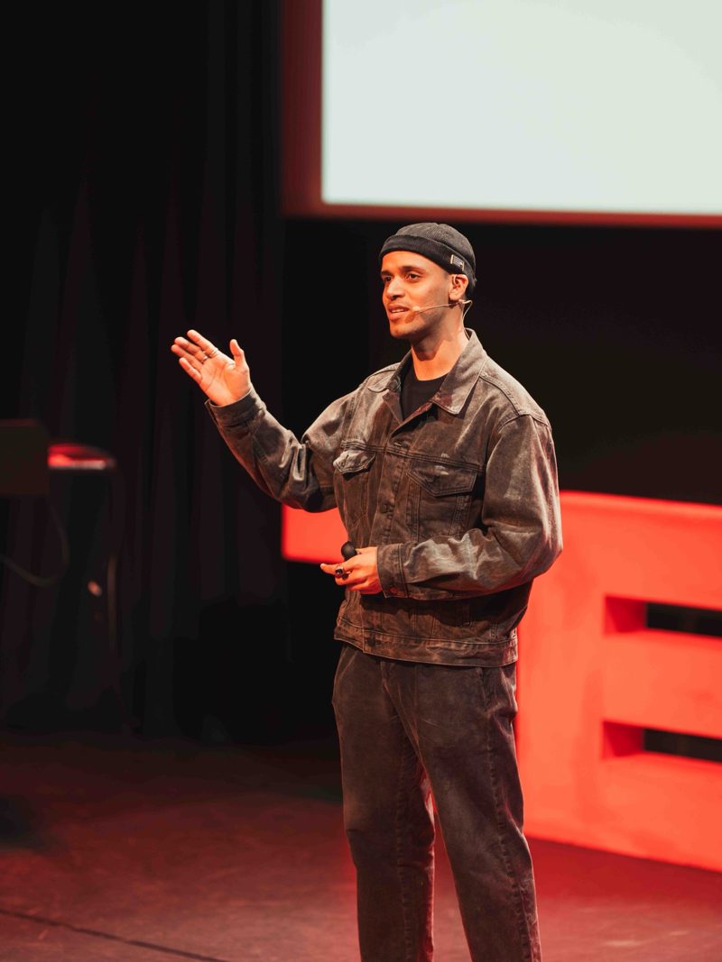 Manoj on stage at TEDxVUAmsterdam, mid-sentence, one hand raised